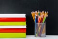 A stack of colorful books and a glass of pencils on the table, on the background of a black school Board