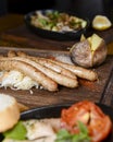 Stack of cooked sausages served on a rustic wooden board with sauce and baked potatoes.