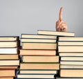 stack of different books on a gray background, hand sticks out from behind a pile and shows up the index finger
