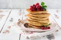 Stack of homemade little pancakes with honey, fresh raspberries and red currants on an old light wooden background.