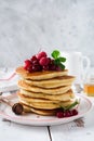 Stack of homemade little pancakes with honey, fresh raspberries and red currants on an old light wooden background.