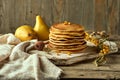 A stack of homemade pancakes on a light wooden board, two pears in the background, a sprig of sea buckthorn and a spoon for honey