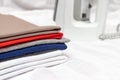 A stack of multicolored clothes close up with the white modern electrical iron of background - ironing, laundry and housework Royalty Free Stock Photo