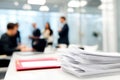 A stack of papers in focus on a desk contrasts with a blurred background of professionals engaged in discussion within a modern Royalty Free Stock Photo