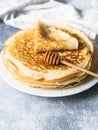 Stack Russian homemade yeast pancakes on white plate, wood honey spoon on grey table. Traditional wheat crepes for Shrovetide