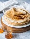 Stack Russian homemade yeast pancakes on white plate, wood honey spoon and honey in jars on grey table. Traditional wheat pancakes