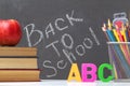 A stack of textbooks, an apple, letters of the alphabet A, B, C, pencils, brushes, paints against a black chalkboard background.