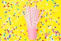 Stacked drinking paper cup striped straws colorful confetti scattered on bright yellow background. Flat lay composition birthday