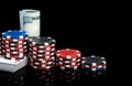 Stacks of poker chips stand on a deck of playing cards next to red cubes and a pack of hundred dollar bills on a glossy black