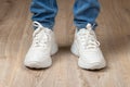 Standing feet shod in white lace-up chunky sole sneakers and blue jeans on the brown floor. Pair of new comfortable shoes for