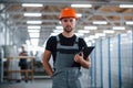 Stands in the corridor with documents in hands. Industrial worker indoors in factory. Young technician with orange hard hat