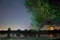 Star tracks in clear deep blue night sky, beautiful countryside landscape with a small lake, willow and rising moon Royalty Free Stock Photo