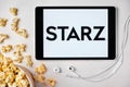 Starz logo on the tablet screen laying on the white table with scattered popcorn and Apple earphones. Spending free time