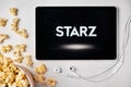 Starz logo on the tablet screen laying on the white table with scattered popcorn and Apple earphones. Spending free time