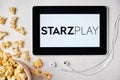 Starzplay logo on the tablet screen laying on the white table with scattered popcorn and Apple earphones. Spending free