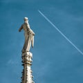 Statue on spire of Milan Cathedral closeup on blue sky background, Milan, Italy. Detail of Gothic roof overlooking flying plane Royalty Free Stock Photo