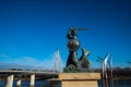 Statue of the Warsaw Mermaid on the banks of the river with a bridge in the background