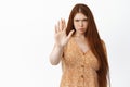 Stay back. Serious young redhead woman extend one hand to stop you, showing prohibit gesture, standing over white