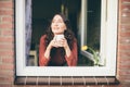 Young smiling woman looking out of window, holding a cup of tea or coffee Royalty Free Stock Photo