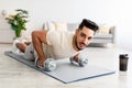 Stay at home fitness. Strong young Arab man doing whole body workout, standing in plank pose, using dumbbells indoors Royalty Free Stock Photo