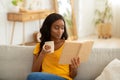 Stay home hobbies concept. Attractive millennial black lady reading book with cup of hot beverage in living room