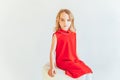 Stay Home Stay Safe. Sweet little girl in red dress sitting on chair against white wall at home, relaxing in white bright living Royalty Free Stock Photo