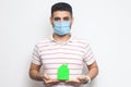 Stay at home, stay safe. Positive man with surgical medical mask holding green small house, looking at camera kindly, reassuring