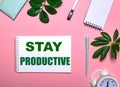 STAY PRODUCTIVE is written in green on a white notepad on a pink background surrounded by notepads, pens, white alarm clock and Royalty Free Stock Photo