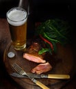 Steak with herbs, hot peppers, cold frothy beer in a glass, vintage knife and fork on a wooden background