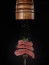 Steak menu. Milled spices falling from pepper mill on grilled pieces of beef steak medium rare on fork on black background