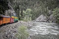 Steam engine rounding a curve with mountain cliff on one side and rushing river on other in pine forested mountains - Vintage