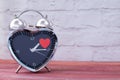 Steel alarm clock in the form of a heart, with a red heart on a pink wooden table and with a white brick wall. Valentine`s Day