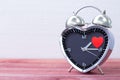 Steel alarm clock in the form of a heart, with a red heart on a pink wooden table and with a white brick wall. Valentine`s Day