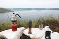 Steel kettle boiling on gas primus, metal mugs and aeropress on cliff on background of lake, brewing coffee at camping. Making hot