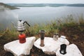 Steel kettle boiling on gas primus, metal mugs and aeropress on cliff on background of lake, brewing coffee at camping. Making hot