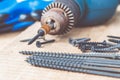 A steel screw is placed on an electric drill near scattered screws on a wooden board background. The concept of tools and repair