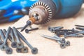 A steel screw is placed on an electric drill near scattered screws on a wooden board background. The concept of tools and repair