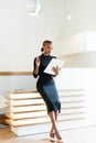 Stern elegant business woman wearing black dress and beige shoes in light office looking at her agenda, full length portrait Royalty Free Stock Photo