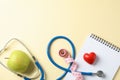 Stethoscope, apple, notebook, heart and measuring tape on yellow background