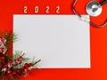 Stethoscope, blank white sheet, wooden number 2022, and fir branches with berries on a red