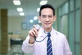 Stethoscope in hand of friendly smiling senior Asian male doctor in white workwear standing in corridor of medical clinic, happy