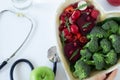 Stethoscope, human hand holding various plant-based food in heart-shape plate, and glass of fresh water on white, top view Royalty Free Stock Photo