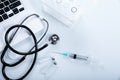 Stethoscope, syringe, vials, and laptop on a white table.