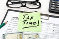 Sticker with phrase `TAX TIME`, tax form, money, pencil, calculator and glasses onoffice table