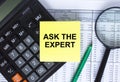 Sticker with text Ask The Expert lying on the calculator. Magnifying glass with green pen on financial documents