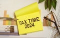 Sticky note with TAX TIME 2024 on financial chart. Taxation and finance reminder concept with pen, glasses, and green plant on Royalty Free Stock Photo