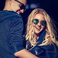 Stilish laughing couple cuddling in modern blue jeans jackets and trendy sunglasses on summer city background. Beautiful female