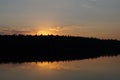 A still dark moody lake at dusk reflects the setting sun on the water surface with a silhouette of trees