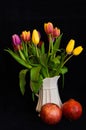 A still life with a bouquet of colourful tulips in a white vase with two pomegranates in front of black background Royalty Free Stock Photo