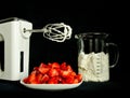 A still life with a bowl of fresh an red strawberries, a food processor, mixer whose wire wisks are full of cream, beside a beaker